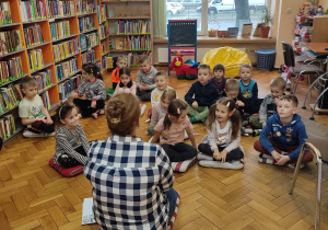 Dzieci siedząc na poduszkach w Bibliotece pośród książek słuchają pilnie opowiadania z serii Przygody Fenka
