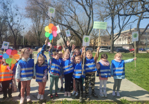 Grupa trzecia ubrana w niebieskie kamizelki odblaskowe uśmiecha się i dzieci z radością trzymają transparenty na temat dbania o środowisko