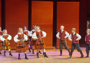Na scenie tańczą dziewczynki i chłopcy w czerwonych strojach