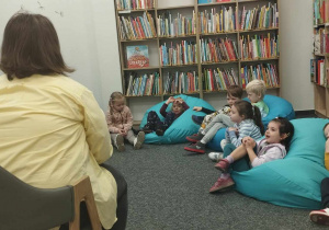 Dzieci leżą na pufach i słuchają opowiadania czytanego przez Panią bibliotekarkę