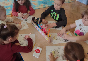 Dzieci przy stoliku kolorują postać. Na środku stołu jest sorter z kredkami