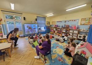 Na zdjęciu dzieci siedzą na kolorowym dywanie w sali przedszkolnej, uważnie oglądając prezentację o psach wyświetlaną na tablicy multimedialnej. Nauczycielka prowadzi zajęcia w ramach „Psiej lekcji”, podczas których dzieci uczą się, jak właściwie opiekować się zwierzętami i jak zachowywać się w ich obecności.