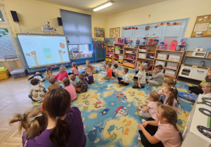 Na zdjęciu dzieci siedzą na dywanie i z zainteresowaniem oglądają prezentację edukacyjną o psach wyświetlaną na tablicy multimedialnej. Podczas „Psiej lekcji” przedszkolaki poznają zasady bezpiecznego kontaktu ze zwierzętami oraz uczą się empatii i odpowiedzialności wobec czworonogów.
