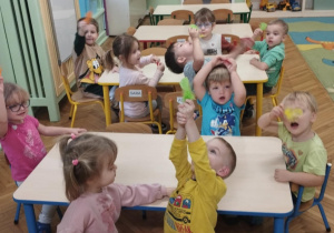 Na zdjęciu dzieci siedzą przy stolikach w sali przedszkolnej i z zaangażowaniem uczestniczą w lekcji oddechowej. Przedszkolaki dmuchają na lekkie piórka, ucząc się kontrolowanego oddechu poprzez zabawę.
