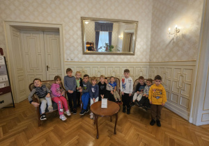Na zdjęciu dzieci siedzą na zabytkowej kanapie we wnętrzu Pałacu Izraela Poznańskiego. Przedszkolaki zwiedzają eleganckie pomieszczenia, poznając historię i wygląd dawnej rezydencji łódzkiego fabrykanta.