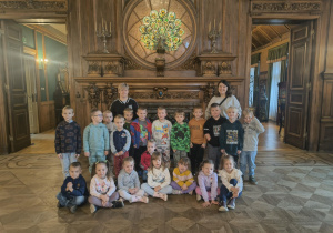 Na zdjęciu grupa przedszkolaków wraz z nauczycielkami pozuje do pamiątkowego zdjęcia we wnętrzu Pałacu Izraela Poznańskiego. Dzieci stoją przed ozdobnym kominkiem i witrażem przedstawiającym pawia, podziwiając piękno zabytkowego wnętrza.