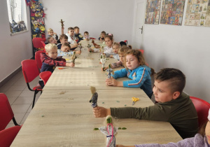 Na zdjęciu dzieci siedzą przy długim stole i z dumą prezentują własnoręcznie wykonane laleczki z trawy i sznurka. Przedszkolaki uczestniczą w warsztatach plastycznych, rozwijając swoją kreatywność i poznając tradycyjne rękodzieło.