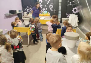Na zdjęciu widać grupę przedszkolaków uczestniczących w zajęciach w sali eksperymentalnej, gdzie prowadząca pokazuje im różne doświadczenia. Dzieci z zaciekawieniem obserwują i aktywnie uczestniczą w warsztatach o tematyce naukowej.
