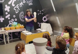 Na zdjęciu prowadząca pokazuje dzieciom eksperyment z balonem, tłumacząc zjawisko w ciekawy i przystępny sposób. Przedszkolaki z zainteresowaniem obserwują doświadczenie, ucząc się poprzez zabawę i odkrywanie.