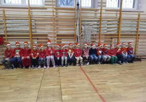 Na zdjęciu widać grupę dzieci siedzących w rzędzie na ławkach w sali gimnastycznej, wszystkie ubrane w czerwone stroje i mikołajkowe czapki. Dzieci patrzą w stronę fotografa, tworząc radosną i świąteczną atmosferę.