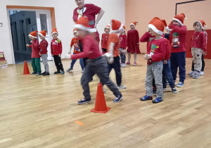 Na zdjęciu dzieci w mikołajkowych czapkach biorą udział w sportowej zabawie na sali gimnastycznej, biegnąc między ustawionymi pachołkami. W tle widać osobę dorosłą, która wspiera i koordynuje przebieg konkurencji.