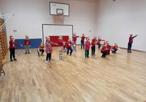 Na zdjęciu dzieci w świątecznych czapkach wykonują ćwiczenia ruchowe na sali gimnastycznej, rozkładając ramiona lub wykonując przysiady. Cała grupa wygląda na zaangażowaną i pełną energii, tworząc radosną, mikołajkową atmosferę.