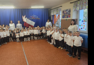 Na zdjęciu widoczna jest duża grupa dzieci ustawionych w półokręgu wraz z kilkorgiem dorosłych, wszyscy odświętnie ubrani i gotowi do wspólnego śpiewania hymnu. W tle znajduje się dekoracja z okazji 11 Listopada, co podkreśla udział przedszkola w akcji „Szkoła do hymnu”.