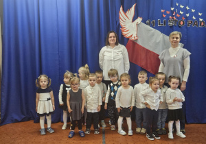 Na zdjęciu widać grupę małych dzieci oraz dwie osoby dorosłe stojące na tle niebieskiej dekoracji z motywami narodowymi i flagą, przygotowaną z okazji 11 Listopada. Uczestnicy ubrane są odświętnie, co podkreśla ich udział w akcji „Szkoła do hymnu”.