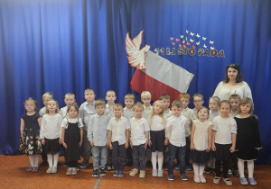 Na zdjęciu znajduje się grupa dzieci ustawionych na tle niebieskiej dekoracji z flagą i symbolem 11 Listopada, wszystkie ubrane odświętnie w jasnych kolorach. Fotografia przedstawia ich udział w akcji „Szkoła do hymnu”, podkreślając uroczysty charakter wydarzenia.