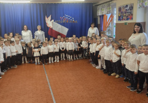 Na zdjęciu widoczna jest duża grupa dzieci ustawionych w półokręgu wraz z kilkorgiem dorosłych, wszyscy odświętnie ubrani i gotowi do wspólnego śpiewania hymnu. W tle znajduje się dekoracja z okazji 11 Listopada, co podkreśla udział przedszkola w akcji „Szkoła do hymnu”.