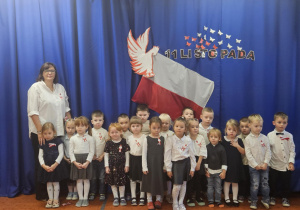 Na zdjęciu widać grupę dzieci stojących na tle niebieskiej dekoracji z flagą i motywem 11 Listopada, wszystkie ubrane odświętnie z biało-czerwonymi akcentami. Fotografia ukazuje ich udział w akcji „Szkoła do hymnu”, podkreślając uroczysty, patriotyczny charakter wydarzenia.