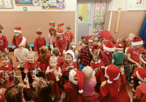 Grupa przedszkolaków w czerwonych strojach i mikołajkowych czapkach bawi się wspólnie w sali przedszkolnej, tworząc radosną, świąteczną atmosferę