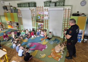 Dzieci siedzą na dywanie i z dużym zainteresowaniem słuchają strażnika, który opowiada o swojej pracy oraz pokazuje im spokojnego, wyszkolonego psa