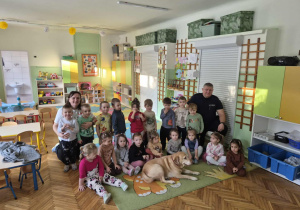 Na zdjęciu widać grupę przedszkolaków siedzących i stojących w sali wraz z nauczycielami oraz spokojnie leżącym psem, podczas wspólnego, radosnego spotkania w przedszkolu