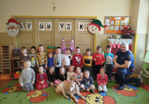 Grupa przedszkolaków pozuje do pamiątkowego zdjęcia w sali, razem ze strażnikiem i spokojnie leżącym psem, tworząc miłą i radosną atmosferę spotkania