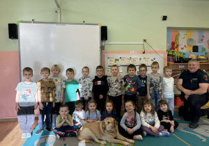 Grupa przedszkolaków ustawia się do wspólnego zdjęcia w sali, a przed nimi spokojnie leży pies, który towarzyszył dzieciom podczas wyjątkowego spotkania z gościem.