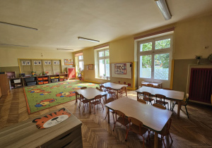 Na zdjęciu widać przestronną i słoneczną salę przedszkolną z dużym, zielonym dywanem w kolorowe postacie. Ustawione stoliki i kąciki zabaw tworzą przyjazną, uporządkowaną przestrzeń sprzyjającą nauce i wspólnej zabawie dzieci.