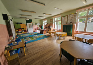 Na zdjęciu znajduje się jasna i przestronna sala przedszkolna z kolorowymi meblami, stolikami oraz dywanem do wspólnej zabawy. Pomieszczenie jest przytulne i dobrze wyposażone, tworząc przyjazne warunki do nauki i aktywności dzieci.