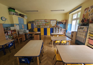 Na zdjęciu widać dobrze zorganizowaną salę przedszkolną z kolorowymi szafkami, stolikami i pomocami dydaktycznymi. Przestrzeń jest jasna i uporządkowana, sprzyjając nauce, zabawie i rozwijaniu samodzielności dzieci.