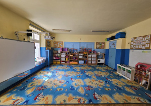 Na zdjęciu widać przestronną salę przedszkolną z dużym, kolorowym dywanem w motyw mapy i statków. Wnętrze jest jasne, pełne zabawek i pomocy dydaktycznych, co tworzy przyjazne miejsce do nauki i zabawy.