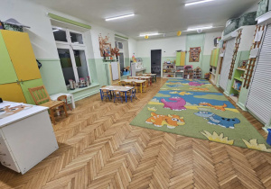 Na zdjęciu znajduje się kolorowa sala przedszkolna z dywanem w dinozaury oraz stolikami dostosowanymi do dzieci. Wnętrze jest jasne, przytulne i dobrze zorganizowane, sprzyjające zabawie oraz nauce w radosnej atmosferze.