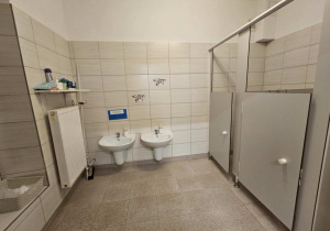 Na zdjęciu widać czystą i zadbaną łazienkę przedszkolną z umywalkami i niskimi kabinami toaletowymi dostosowanymi do potrzeb dzieci. Jasne płytki i proste wyposażenie tworzą przyjazne i bezpieczne miejsce do codziennej higieny.